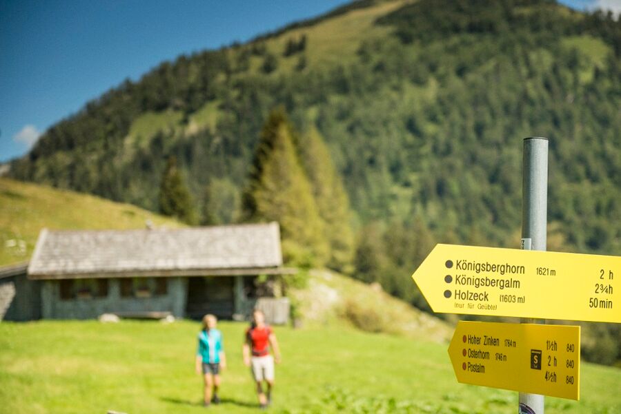 Zwei junge Menschen beim Wandern. Hütte im Hintergrund. Wandertafel im Vordergrund.